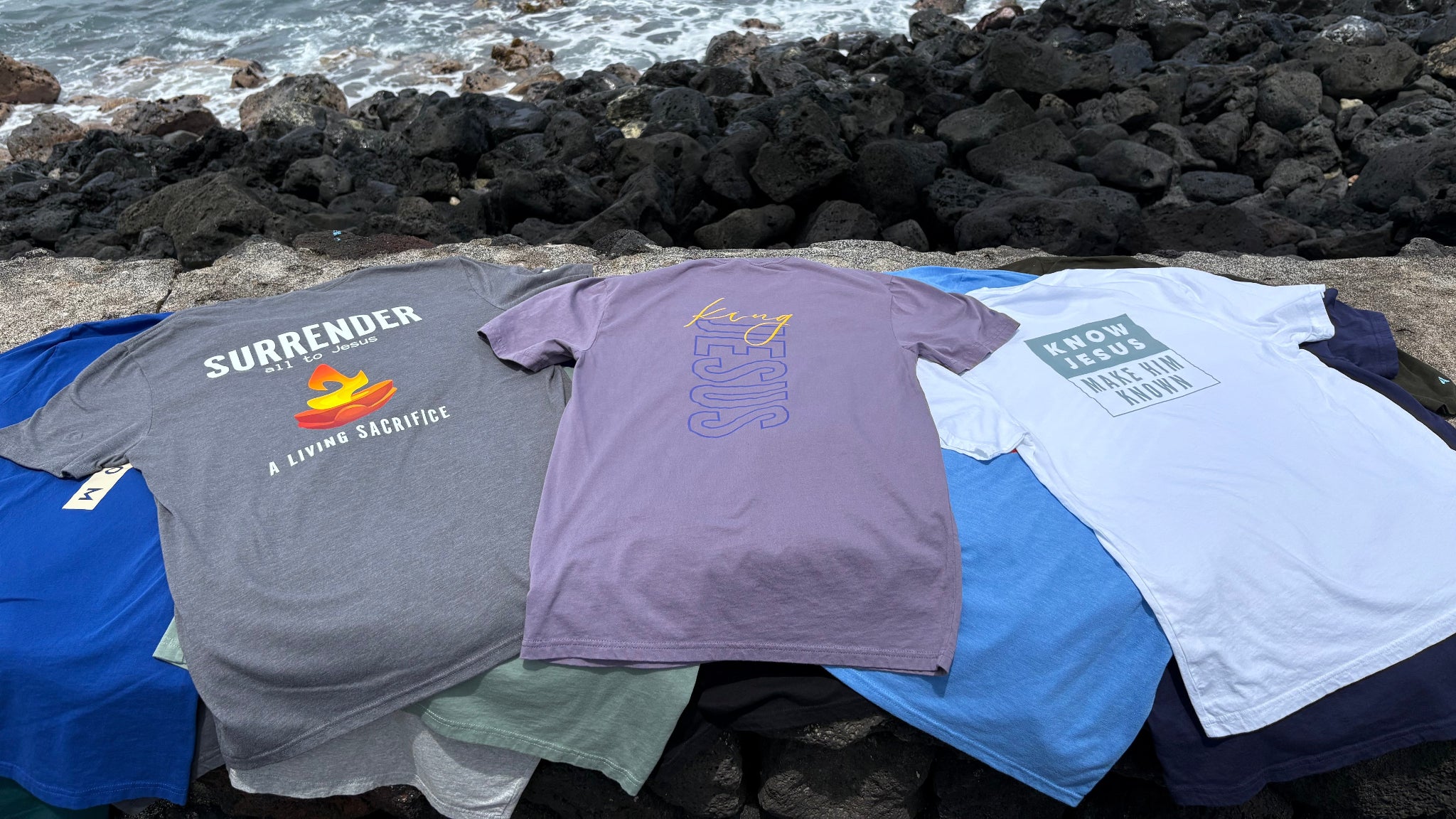 Four t-shirts laid out on a stone ledge with ocean waves in the background
