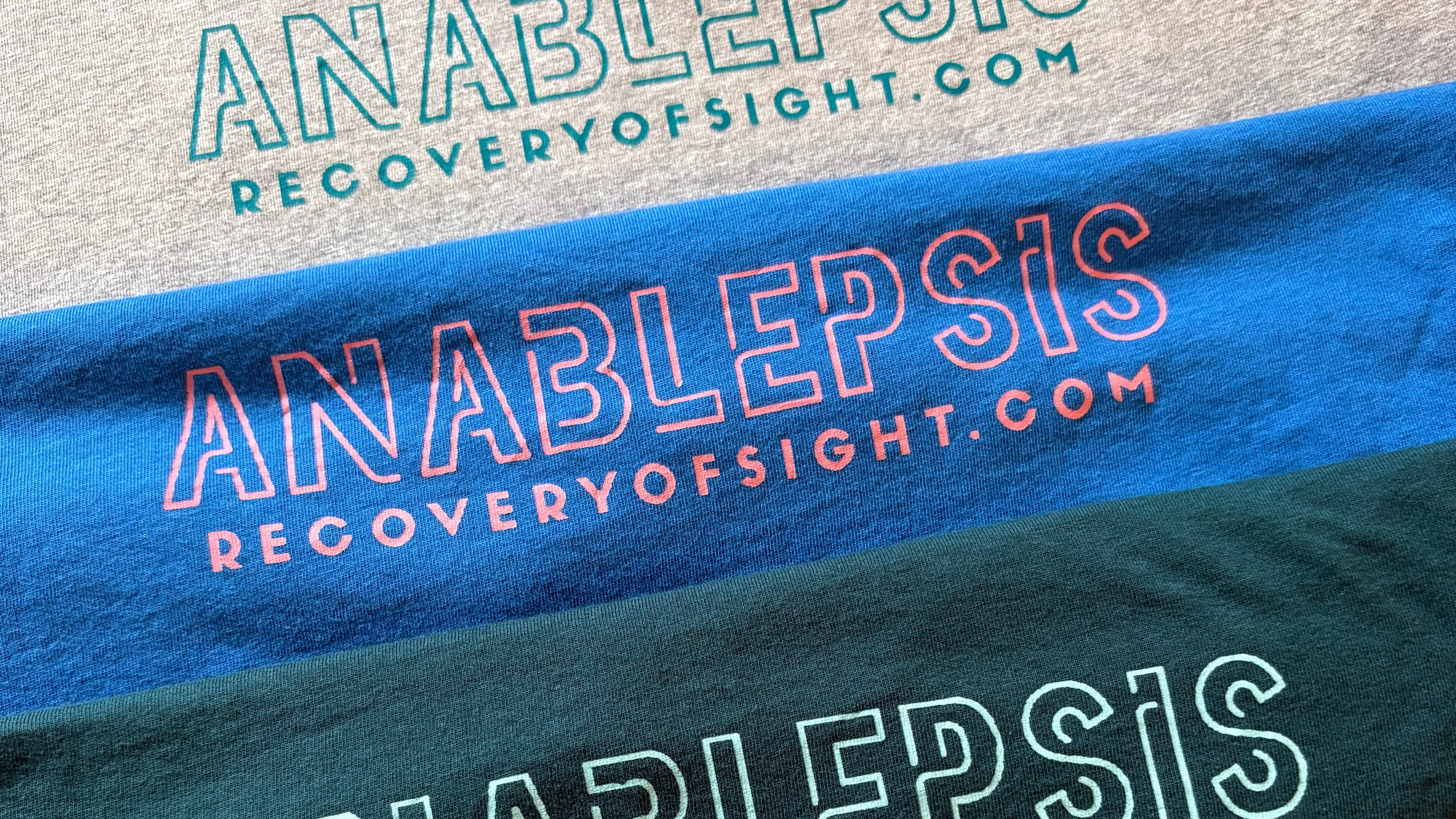 Four t-shirts with 'Anablepsis' and 'recoveryofsight.com' text on a textured surface.